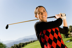 woman in a red and black diamond sweater smiling as she hits a golf ball.